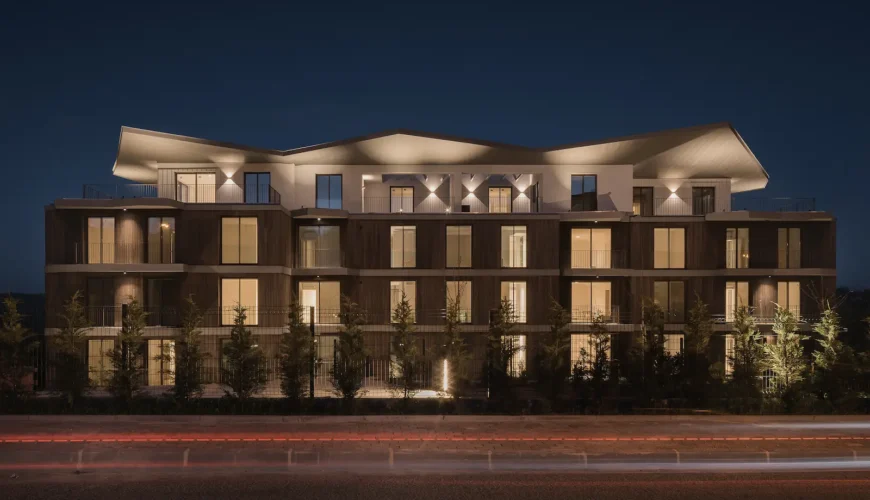 Edifício de apartamentos moderno de quatro andares com revestimento em madeira escura e grandes janelas. O telhado é cinzento e possui uma forma geométrica ondulada proeminente, saliente. O edifício apresenta varandas em toda a fachada. Em primeiro plano, há um jardim relvado bem cuidado com pequenos arbustos e árvores jovens, ladeado por uma rampa de betão.