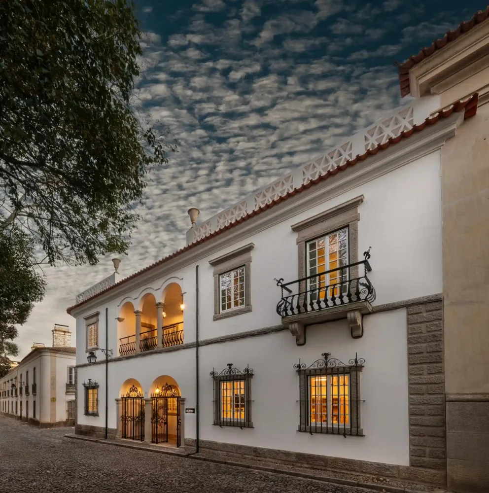 Vista exterior de um edifício tradicional português (possivelmente um palacete ou solar) com paredes brancas, telhado de telhas e detalhes em pedra. O edifício apresenta janelas com grades de ferro forjado iluminadas e uma varanda no primeiro andar com balaustrada em ferro. A entrada principal tem um portão de ferro sob uma arcada. A rua é de calçada portuguesa e o céu está dramático com nuvens.
