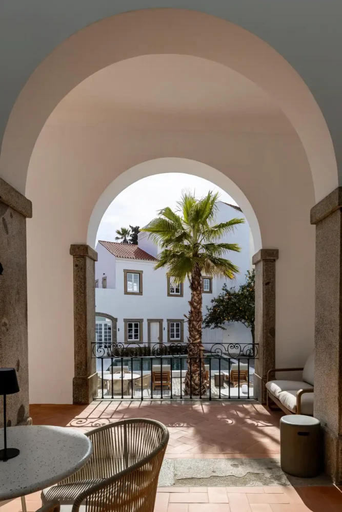 Vista do pátio e da piscina enquadrada por um grande arco branco sustentado por pilares de pedra rústica. Dentro do arco, vê-se uma palmeira, a piscina e o exterior branco do edifício. Em primeiro plano, uma área de estar sombreada com uma mesa redonda e cadeiras de vime, criando uma sensação de retiro acolhedor.