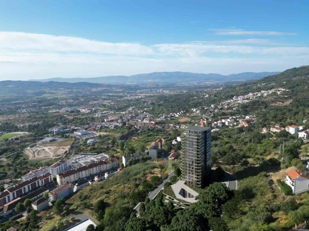 Uma vista aérea da Green View Tower / Torre de Santo António na Covilhã alto e moderno no topo de uma colina arborizada, com uma ampla paisagem de vale e montanhas no fundo, sob um céu azul claro com algumas nuvens brancas. O edifício está rodeado por vegetação e uma rampa de acesso serpenteante. A área urbana circundante é visível, misturando-se com a paisagem natural.