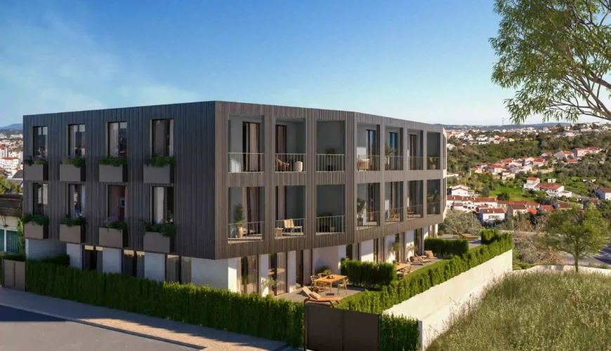 Renderização exterior, vista de ângulo, de um moderno edifício de apartamentos de três andares. A fachada é revestida com ripas escuras e tem varandas e pátios no rés-do-chão com vista para uma área urbana.