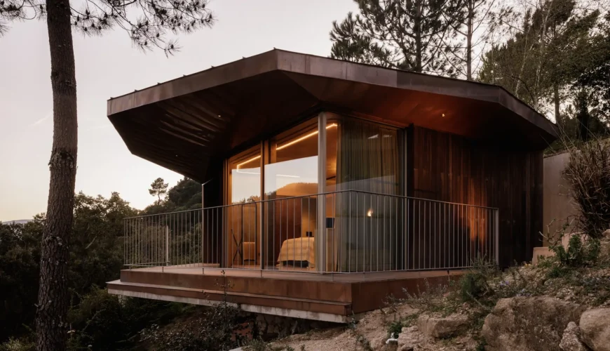 Vista exterior da Douro Wood House com design hexagonal ou curvo, revestido a madeira escura. Uma grande varanda suspensa tem vista para a paisagem arborizada. As janelas iluminadas mostram o interior acolhedor.
